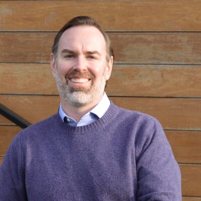 Portrait of man with short brown hair, smiling in a purple sweater in front of a paneled wood background