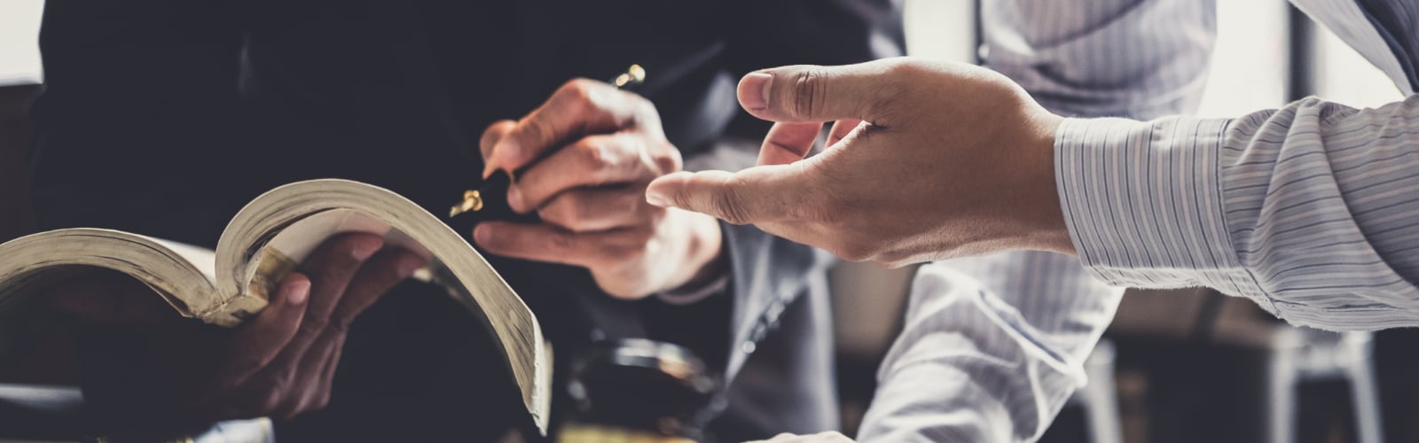 closeup of two people hands, one holding a book and pen, the other pointing at the book