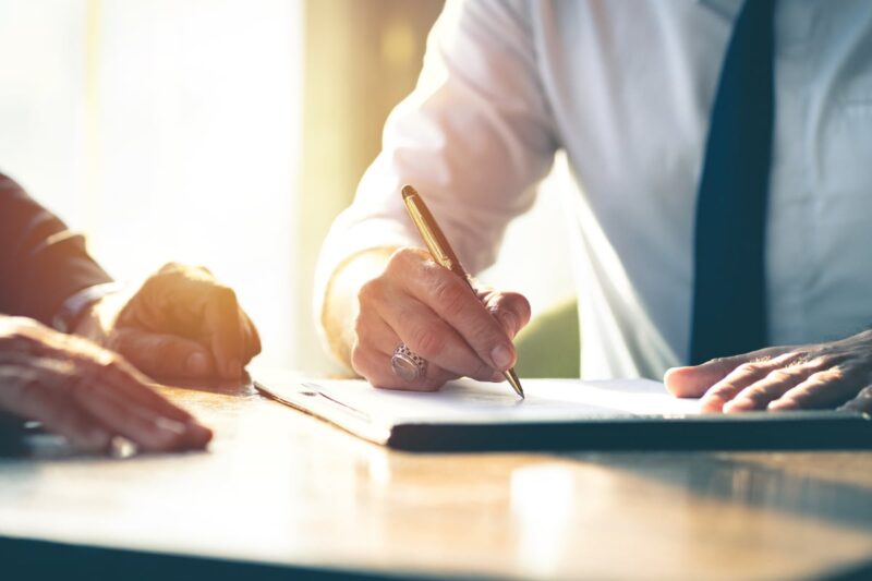 Hand with a pen signing a document on a desk and another set of hands resting on desk